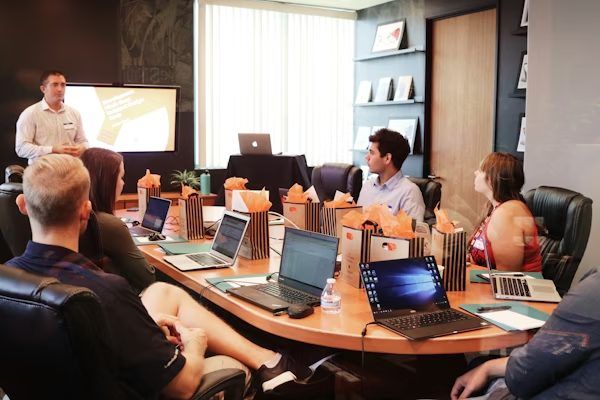 Team sitzt mit Laptops an einem langen Besprechungstisch und hört einer Präsentation zu